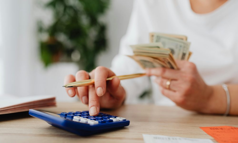 A person sits at a desk calculating finances using a calculator and holding cash.