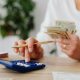 A person sits at a desk calculating finances using a calculator and holding cash.